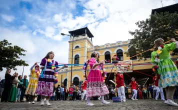 Dia do Manezinho celebra cultura e tradições de Florianópolis Participantes vestidos com trajes coloridos realizam uma apresentação cultural em frente ao Mercado Público de Florianópolis durante uma celebração do Dia do Manezinho. A imagem ilustra a valorização das tradições, da cultura popular e das manifestações folclóricas que fazem parte da identidade da cidade.