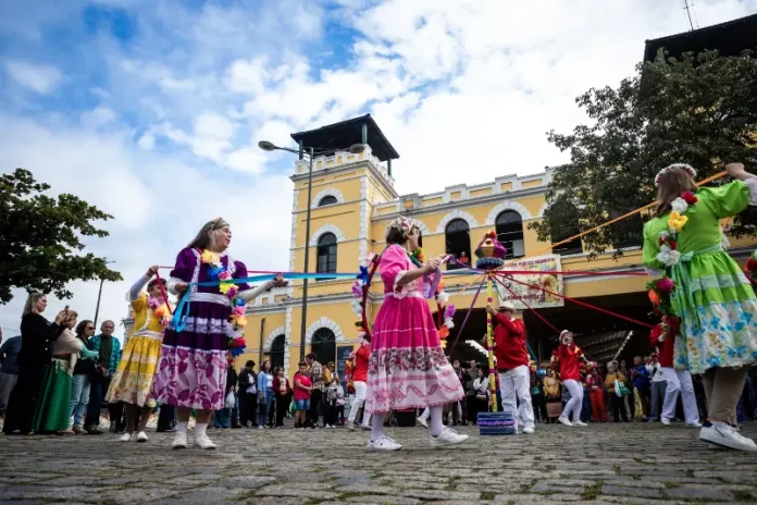 dia-do-manezinho Participantes vestidos com trajes coloridos realizam uma apresentação cultural em frente ao Mercado Público de Florianópolis durante uma celebração do Dia do Manezinho. A imagem ilustra a valorização das tradições, da cultura popular e das manifestações folclóricas que fazem parte da identidade da cidade.