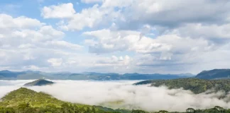 Vista panorâmica de montanhas e áreas de vegetação em Santa Catarina, com neblina cobrindo os vales e uma estrada atravessando a paisagem sob céu com nuvens. A imagem ilustra a atuação de uma massa de ar frio no estado, responsável pela manutenção do tempo firme e pela queda das temperaturas ao longo da semana.