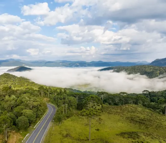 Vista panorâmica de montanhas e áreas de vegetação em Santa Catarina, com neblina cobrindo os vales e uma estrada atravessando a paisagem sob céu com nuvens. A imagem ilustra a atuação de uma massa de ar frio no estado, responsável pela manutenção do tempo firme e pela queda das temperaturas ao longo da semana.