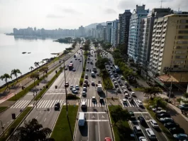 Florianópolis envia à Câmara projeto de nova regulamentação do transporte por aplicativos Vista aérea de uma avenida à beira-mar em Florianópolis com intenso fluxo de veículos e prédios residenciais ao lado. A imagem ilustra o contexto da mobilidade urbana na Capital e acompanha matéria sobre projeto de lei que atualiza as regras do transporte por aplicativos para ampliar segurança, fiscalização e qualidade do serviço.
