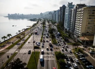 Florianópolis envia à Câmara projeto de nova regulamentação do transporte por aplicativos Vista aérea de uma avenida à beira-mar em Florianópolis com intenso fluxo de veículos e prédios residenciais ao lado. A imagem ilustra o contexto da mobilidade urbana na Capital e acompanha matéria sobre projeto de lei que atualiza as regras do transporte por aplicativos para ampliar segurança, fiscalização e qualidade do serviço.