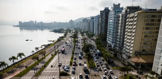 Florianópolis envia à Câmara projeto de nova regulamentação do transporte por aplicativos Vista aérea de uma avenida à beira-mar em Florianópolis com intenso fluxo de veículos e prédios residenciais ao lado. A imagem ilustra o contexto da mobilidade urbana na Capital e acompanha matéria sobre projeto de lei que atualiza as regras do transporte por aplicativos para ampliar segurança, fiscalização e qualidade do serviço.