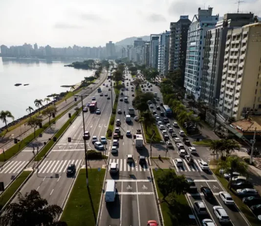 Florianópolis envia à Câmara projeto de nova regulamentação do transporte por aplicativos Vista aérea de uma avenida à beira-mar em Florianópolis com intenso fluxo de veículos e prédios residenciais ao lado. A imagem ilustra o contexto da mobilidade urbana na Capital e acompanha matéria sobre projeto de lei que atualiza as regras do transporte por aplicativos para ampliar segurança, fiscalização e qualidade do serviço.
