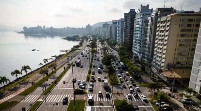 Vista aérea de uma avenida à beira-mar em Florianópolis com intenso fluxo de veículos e prédios residenciais ao lado. A imagem ilustra o contexto da mobilidade urbana na Capital e acompanha matéria sobre projeto de lei que atualiza as regras do transporte por aplicativos para ampliar segurança, fiscalização e qualidade do serviço.