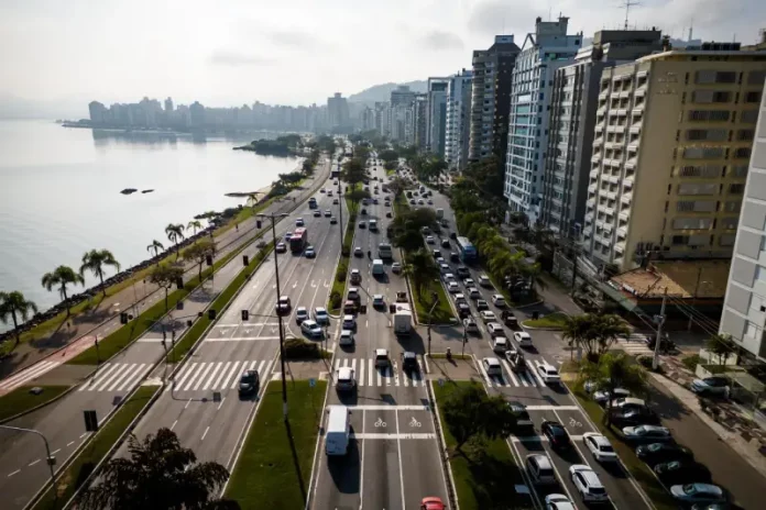 transporte por aplicativos Vista aérea de uma avenida à beira-mar em Florianópolis com intenso fluxo de veículos e prédios residenciais ao lado. A imagem ilustra o contexto da mobilidade urbana na Capital e acompanha matéria sobre projeto de lei que atualiza as regras do transporte por aplicativos para ampliar segurança, fiscalização e qualidade do serviço.