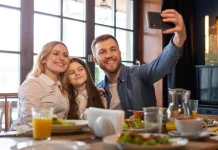 Dia dos Pais deve impulsionar vendas em bares e restaurantes Foto de pai, mãe e filha, em restaurante, fazendo uma selfie, simbolizando almoço em família no Dia dos Pais