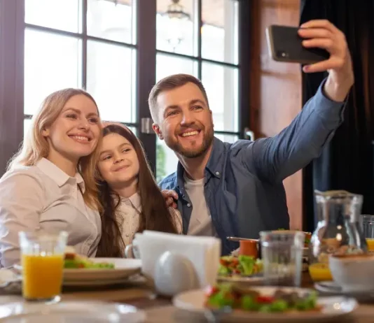 Dia dos Pais deve impulsionar vendas em bares e restaurantes Foto de pai, mãe e filha, em restaurante, fazendo uma selfie, simbolizando almoço em família no Dia dos Pais