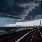 Frente fria provoca chuva e aumenta o risco de temporais em Santa Catarina A imagem mostra uma frente fria sobre o litoral de Santa Catarina. O céu está coberto por nuvens escuras e densas, enquanto um raio cai próximo ao mar. Em primeiro plano, aparecem uma ciclovia e uma avenida à beira-mar, com postes de iluminação ao longo do percurso. Ao fundo, montanhas surgem sob a forte instabilidade, reforçando o cenário de chuva e temporais.