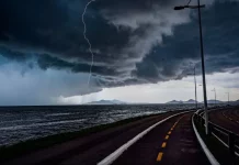 Frente fria provoca chuva e aumenta o risco de temporais em Santa Catarina A imagem mostra uma frente fria sobre o litoral de Santa Catarina. O céu está coberto por nuvens escuras e densas, enquanto um raio cai próximo ao mar. Em primeiro plano, aparecem uma ciclovia e uma avenida à beira-mar, com postes de iluminação ao longo do percurso. Ao fundo, montanhas surgem sob a forte instabilidade, reforçando o cenário de chuva e temporais.