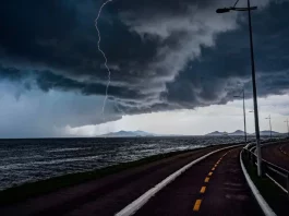 Frente fria provoca chuva e aumenta o risco de temporais em Santa Catarina A imagem mostra uma frente fria sobre o litoral de Santa Catarina. O céu está coberto por nuvens escuras e densas, enquanto um raio cai próximo ao mar. Em primeiro plano, aparecem uma ciclovia e uma avenida à beira-mar, com postes de iluminação ao longo do percurso. Ao fundo, montanhas surgem sob a forte instabilidade, reforçando o cenário de chuva e temporais.