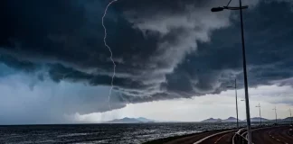 Frente fria provoca chuva e aumenta o risco de temporais em Santa Catarina A imagem mostra uma frente fria sobre o litoral de Santa Catarina. O céu está coberto por nuvens escuras e densas, enquanto um raio cai próximo ao mar. Em primeiro plano, aparecem uma ciclovia e uma avenida à beira-mar, com postes de iluminação ao longo do percurso. Ao fundo, montanhas surgem sob a forte instabilidade, reforçando o cenário de chuva e temporais.