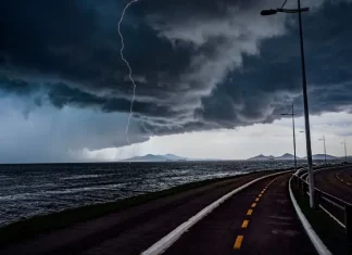 Frente fria provoca chuva e aumenta o risco de temporais em Santa Catarina A imagem mostra uma frente fria sobre o litoral de Santa Catarina. O céu está coberto por nuvens escuras e densas, enquanto um raio cai próximo ao mar. Em primeiro plano, aparecem uma ciclovia e uma avenida à beira-mar, com postes de iluminação ao longo do percurso. Ao fundo, montanhas surgem sob a forte instabilidade, reforçando o cenário de chuva e temporais.