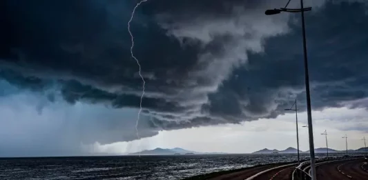Frente fria provoca chuva e aumenta o risco de temporais em Santa Catarina A imagem mostra uma frente fria sobre o litoral de Santa Catarina. O céu está coberto por nuvens escuras e densas, enquanto um raio cai próximo ao mar. Em primeiro plano, aparecem uma ciclovia e uma avenida à beira-mar, com postes de iluminação ao longo do percurso. Ao fundo, montanhas surgem sob a forte instabilidade, reforçando o cenário de chuva e temporais.