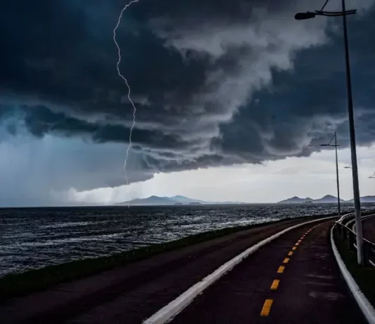 Frente fria provoca chuva e aumenta o risco de temporais em Santa Catarina A imagem mostra uma frente fria sobre o litoral de Santa Catarina. O céu está coberto por nuvens escuras e densas, enquanto um raio cai próximo ao mar. Em primeiro plano, aparecem uma ciclovia e uma avenida à beira-mar, com postes de iluminação ao longo do percurso. Ao fundo, montanhas surgem sob a forte instabilidade, reforçando o cenário de chuva e temporais.
