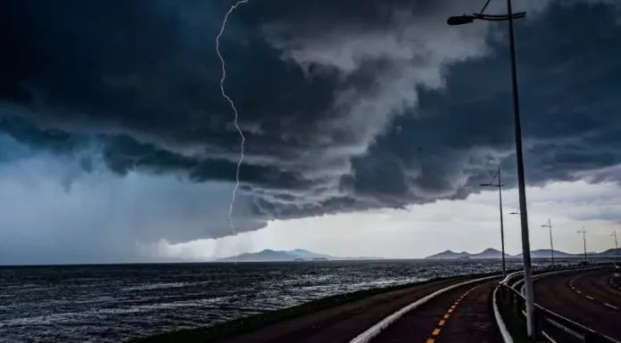 Frente fria provoca chuva e aumenta o risco de temporais em Santa Catarina A imagem mostra uma frente fria sobre o litoral de Santa Catarina. O céu está coberto por nuvens escuras e densas, enquanto um raio cai próximo ao mar. Em primeiro plano, aparecem uma ciclovia e uma avenida à beira-mar, com postes de iluminação ao longo do percurso. Ao fundo, montanhas surgem sob a forte instabilidade, reforçando o cenário de chuva e temporais.