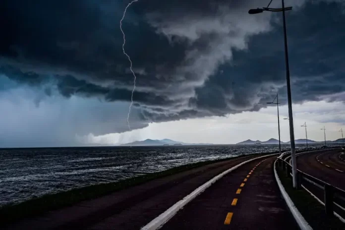 A imagem mostra uma frente fria sobre o litoral de Santa Catarina. O céu está coberto por nuvens escuras e densas, enquanto um raio cai próximo ao mar. Em primeiro plano, aparecem uma ciclovia e uma avenida à beira-mar, com postes de iluminação ao longo do percurso. Ao fundo, montanhas surgem sob a forte instabilidade, reforçando o cenário de chuva e temporais.