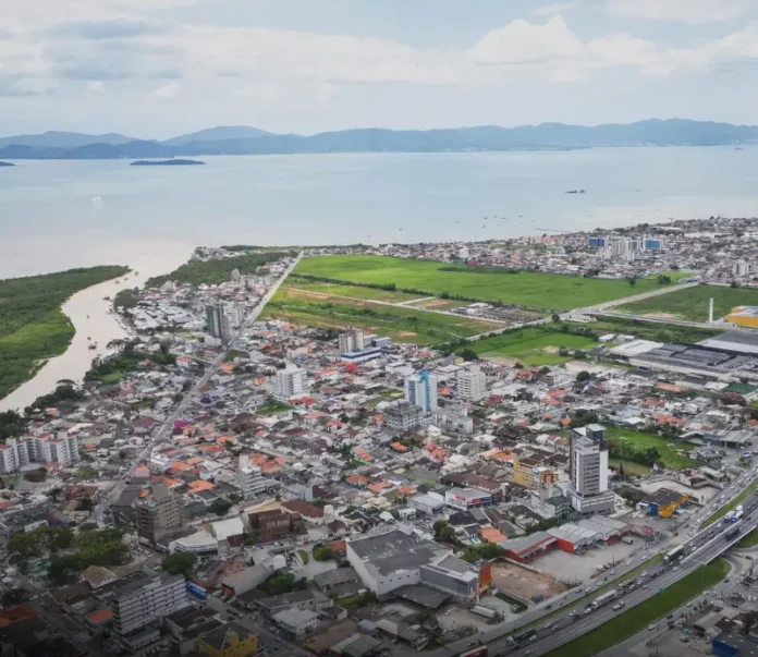 Vista aérea de Biguaçu mostra a área urbana do município, com prédios, casas, vias e a Baía Norte ao fundo, ilustrando a cidade que chegou a 85.472 habitantes em 2026.