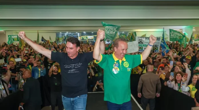 Flávio Bolsonaro e Jorginho Mello aparecem de mãos levantadas diante de apoiadores durante ato de campanha em Santa Catarina.