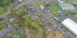 Chuvas e temporais provocam alagamentos em área urbana, com água barrenta atingindo terrenos e residências próximas a uma via.