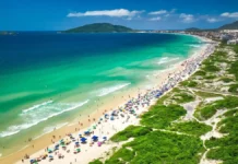 Vista aérea de praia movimentada em Florianópolis, com banhistas, guarda-sóis e área de circulação onde atuam comerciantes ambulantes durante o verão.