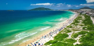 Vista aérea de praia movimentada em Florianópolis, com banhistas, guarda-sóis e área de circulação onde atuam comerciantes ambulantes durante o verão.