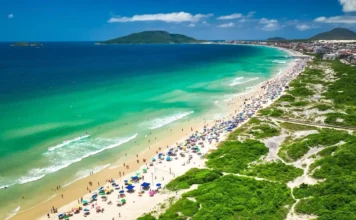 Carrinhos de ambulantes terão QR Code e microchip nas praias de Florianópolis Vista aérea de praia movimentada em Florianópolis, com banhistas, guarda-sóis e área de circulação onde atuam comerciantes ambulantes durante o verão.