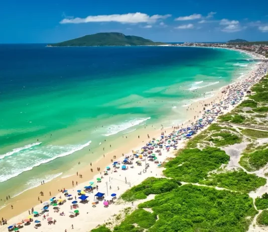 Carrinhos de ambulantes terão QR Code e microchip nas praias de Florianópolis Vista aérea de praia movimentada em Florianópolis, com banhistas, guarda-sóis e área de circulação onde atuam comerciantes ambulantes durante o verão.