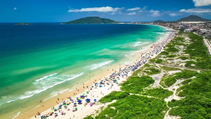 Vista aérea de praia movimentada em Florianópolis, com banhistas, guarda-sóis e área de circulação onde atuam comerciantes ambulantes durante o verão.