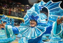 Integrante de escola de samba desfila com fantasia azul brilhante e braços abertos, diante do público, durante o Carnaval de Florianópolis. A imagem ilustra a programação do Carnaval 2027 na Capital.