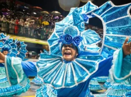 Integrante de escola de samba desfila com fantasia azul brilhante e braços abertos, diante do público, durante o Carnaval de Florianópolis. A imagem ilustra a programação do Carnaval 2027 na Capital.