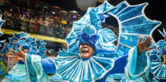 Integrante de escola de samba desfila com fantasia azul brilhante e braços abertos, diante do público, durante o Carnaval de Florianópolis. A imagem ilustra a programação do Carnaval 2027 na Capital.