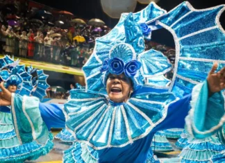 Integrante de escola de samba desfila com fantasia azul brilhante e braços abertos, diante do público, durante o Carnaval de Florianópolis. A imagem ilustra a programação do Carnaval 2027 na Capital.