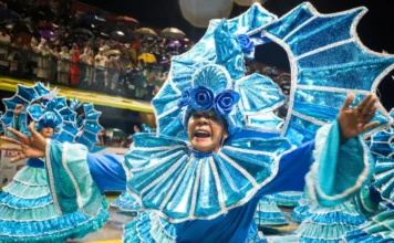 Integrante de escola de samba desfila com fantasia azul brilhante e braços abertos, diante do público, durante o Carnaval de Florianópolis. A imagem ilustra a programação do Carnaval 2027 na Capital.