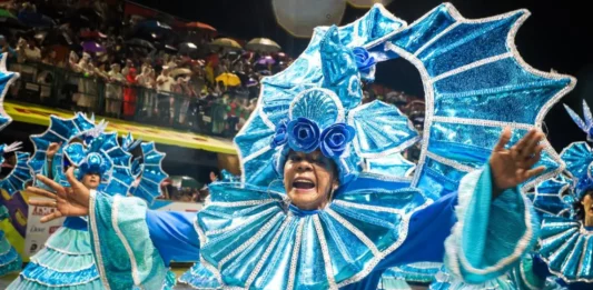 Integrante de escola de samba desfila com fantasia azul brilhante e braços abertos, diante do público, durante o Carnaval de Florianópolis. A imagem ilustra a programação do Carnaval 2027 na Capital.