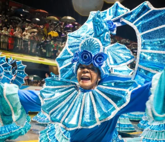 Integrante de escola de samba desfila com fantasia azul brilhante e braços abertos, diante do público, durante o Carnaval de Florianópolis. A imagem ilustra a programação do Carnaval 2027 na Capital.
