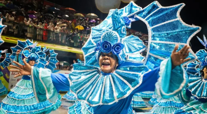 Carnaval 2027 em Florianópolis terá shows gratuitos em nove arenas Integrante de escola de samba desfila com fantasia azul brilhante e braços abertos, diante do público, durante o Carnaval de Florianópolis. A imagem ilustra a programação do Carnaval 2027 na Capital.