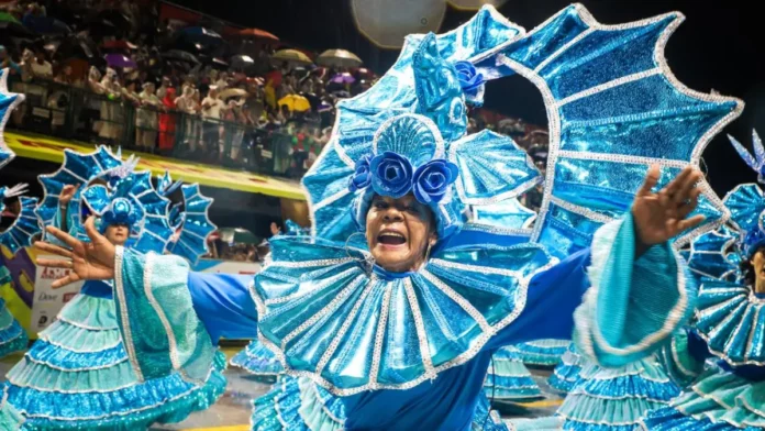 Integrante de escola de samba desfila com fantasia azul brilhante e braços abertos, diante do público, durante o Carnaval de Florianópolis. A imagem ilustra a programação do Carnaval 2027 na Capital.