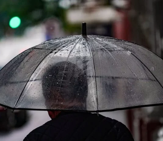 Pessoa caminha com guarda-chuva em rua molhada, ilustrando a previsão de chuva e temporais em Santa Catarina.