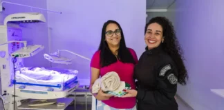 Duas mulheres apresentam kits com toucas e cueiros destinados aos recém-nascidos do Hospital Regional de São José, produzidos por internas do sistema prisional catarinense.