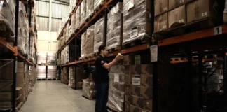 Trabalhador confere produtos armazenados em depósito, ilustrando as compras públicas e o controle de gastos em licitações de Santa Catarina.