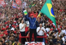 Lula segura a bandeira do Brasil diante de uma multidão de apoiadores em ato político. Presidente foi reeleito para o quarto mandato em 2026.