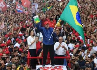 Lula segura a bandeira do Brasil diante de uma multidão de apoiadores em ato político. Presidente foi reeleito para o quarto mandato em 2026.