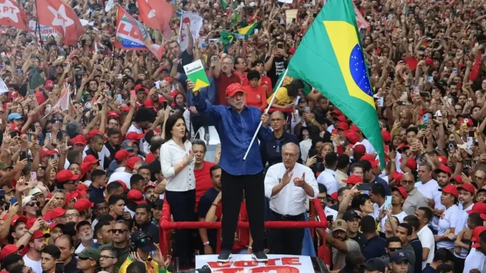 lula-reeleito-1200x675 Lula segura a bandeira do Brasil diante de uma multidão de apoiadores em ato político. Presidente foi reeleito para o quarto mandato em 2026.