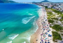Operação Verão 2026/2027: Florianópolis terá Réveillon ampliado e reforço nos serviços Vista aérea de praia movimentada em Florianópolis, com banhistas, guarda-sóis e mar azul, ilustrando a temporada de verão 2026/2027.