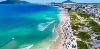 Vista aérea de praia movimentada em Florianópolis, com banhistas, guarda-sóis e mar azul, ilustrando a temporada de verão 2026/2027.