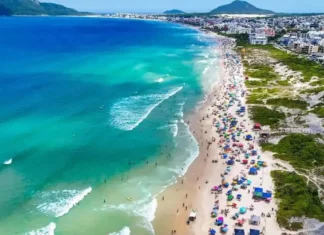 Operação Verão 2026/2027: Florianópolis terá Réveillon ampliado e reforço nos serviços Vista aérea de praia movimentada em Florianópolis, com banhistas, guarda-sóis e mar azul, ilustrando a temporada de verão 2026/2027.
