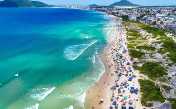 Vista aérea de praia movimentada em Florianópolis, com banhistas, guarda-sóis e mar azul, ilustrando a temporada de verão 2026/2027.