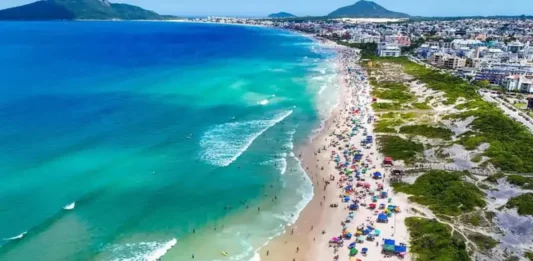 Operação Verão 2026/2027: Florianópolis terá Réveillon ampliado e reforço nos serviços Vista aérea de praia movimentada em Florianópolis, com banhistas, guarda-sóis e mar azul, ilustrando a temporada de verão 2026/2027.