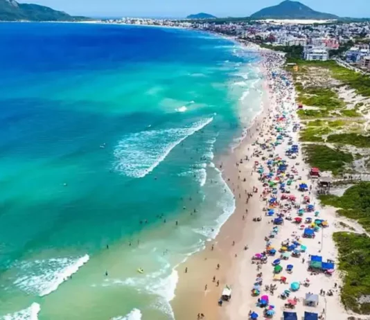 Vista aérea de praia movimentada em Florianópolis, com banhistas, guarda-sóis e mar azul, ilustrando a temporada de verão 2026/2027.